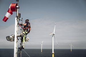 Zu sehen ist ein hochqualifizierter Elektrotechniker bei der Wartung der Kommunikationseinheit eines Offshore Windparks. Im Hintergrund sind Windkraftanlagen zu sehen.